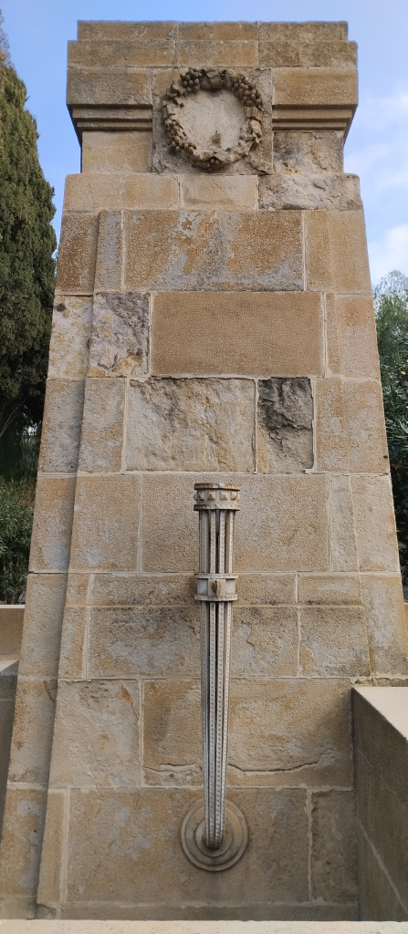 Detall del lateral del pilar monumental, amb decoració escultòrica d'una corona de flors a la part superior i d'una torxa de pedra a la part baixa.